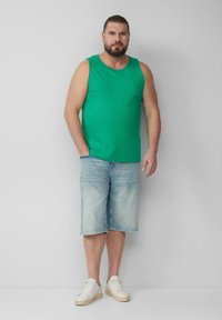 Zielony top na ramiączkach z lekkiego materiału, zestawiony z jasnoniebieskimi szortami dżinsowymi i białymi sneakersami. Modelka stoi z jedną ręką w kieszeni.