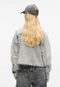 Szara kurtka dżinsowa z surowym dołem, w zestawieniu z czarnymi dżinsami. Model nosi szary kapelusz baseballowy. Włosy są długie i faliste, widok od tyłu.