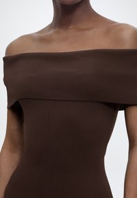 Γυναίκα που φοράει καφέ πλεκτό φόρεμα με ρίγες, χωρίς ώμους, που αποκαλύπτει το λαιμό και το πάνω μέρος του σώματος, μπροστά σε ένα απλό φόντο.