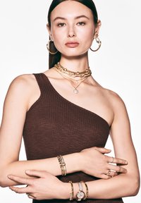 Joyería de oro en capas sobre una modelo, incluyendo pendientes de aro, múltiples collares y varias pulseras, combinados con una blusa marrón de canalé.