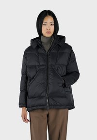 Doudoune noire avec fermeture éclair, capuche amovible, et grandes poches avant. Présente un motif géométrique matelassé et une finition en tissu durable.
