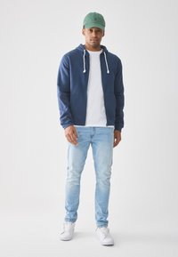 Mann som har på seg grønn caps, marineblå hettejakke, hvit t-skjorte, lyseblå jeans og hvite joggesko, stående mot en hvit bakgrunn.