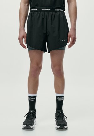 Männlicher Sportler trägt schwarze Laufshorts, schwarze Socken mit dem Text "BORN RUN" und schwarze Laufschuhe, steht mit leicht auseinanderstehenden Beinen.