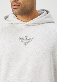 Felpa grigia con una texture morbida, dotata di un emblema in argento in rilievo sul petto. Design semplice con cappuccio regolabile e maniche raglan.