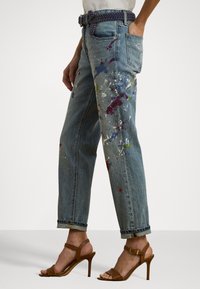 Light blue denim jeans with a loose fit, featuring colorful paint splatters. Paired with brown high-heeled sandals and a woven belt.