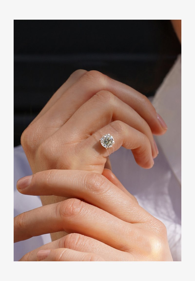Mano con anillo solitario de diamante claro en el dedo anular, sostenida suavemente por la otra mano sobre un fondo neutro.