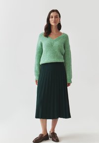 Un pull tricoté vert clair avec un col en V, associé à une jupe midi plissée vert foncé et des chaussures à enfiler marron. Design simple et texturé.