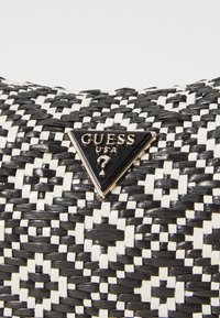 Sac tissé noir et blanc avec un logo triangulaire Guess en émail noir ; la texture met en valeur un motif en croix.