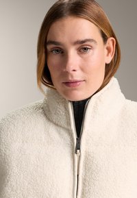 Fleecejacke in Cremefarbe mit hohem Kragen, ausgestattet mit einem Frontreißverschluss und strukturierter Oberfläche. Gefüttertes Innenfutter in kontrastierender Farbe.