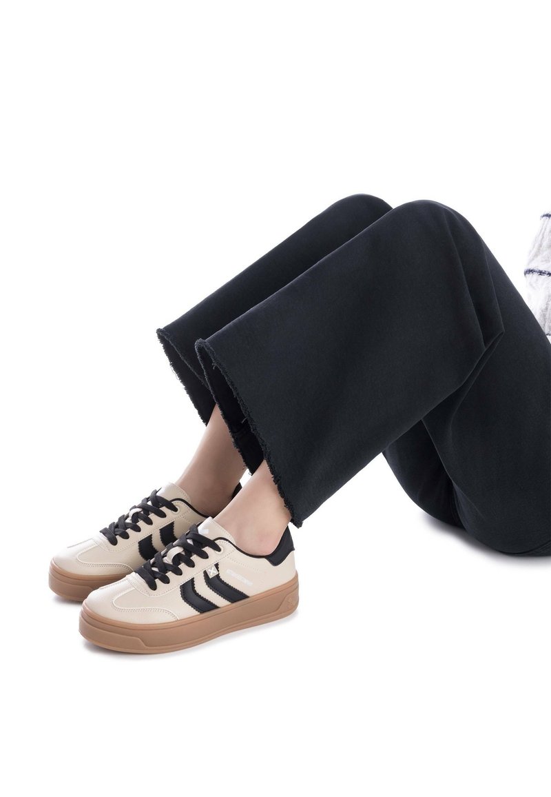 Zapatillas con una parte superior de cuero crema, acentos negros y cordones negros; suela de goma marrón. Llevadas con pantalones anchos negros con dobladillos deshilachados.