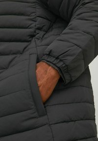 Sort pufferjakke med kviltet design, som har ribbede manchetter og en sidelommer. Teksturen ser isoleret og holdbar ud.