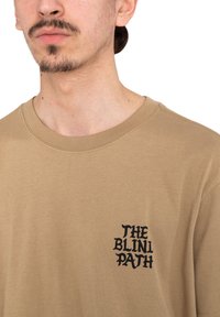 Beige short-sleeve t-shirt featuring a black graphic text logo "THE BLINI PATH" on the left chest. Cotton material with a soft texture.