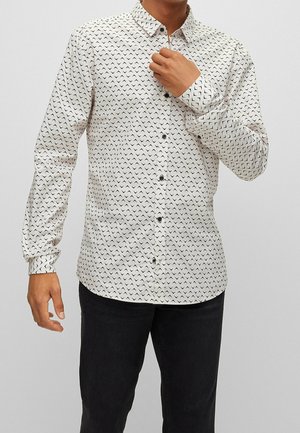 Homme portant une chemise blanche boutonnée avec des motifs géométriques en chevron noirs et un pantalon noir, ajustant le col avec la main droite.