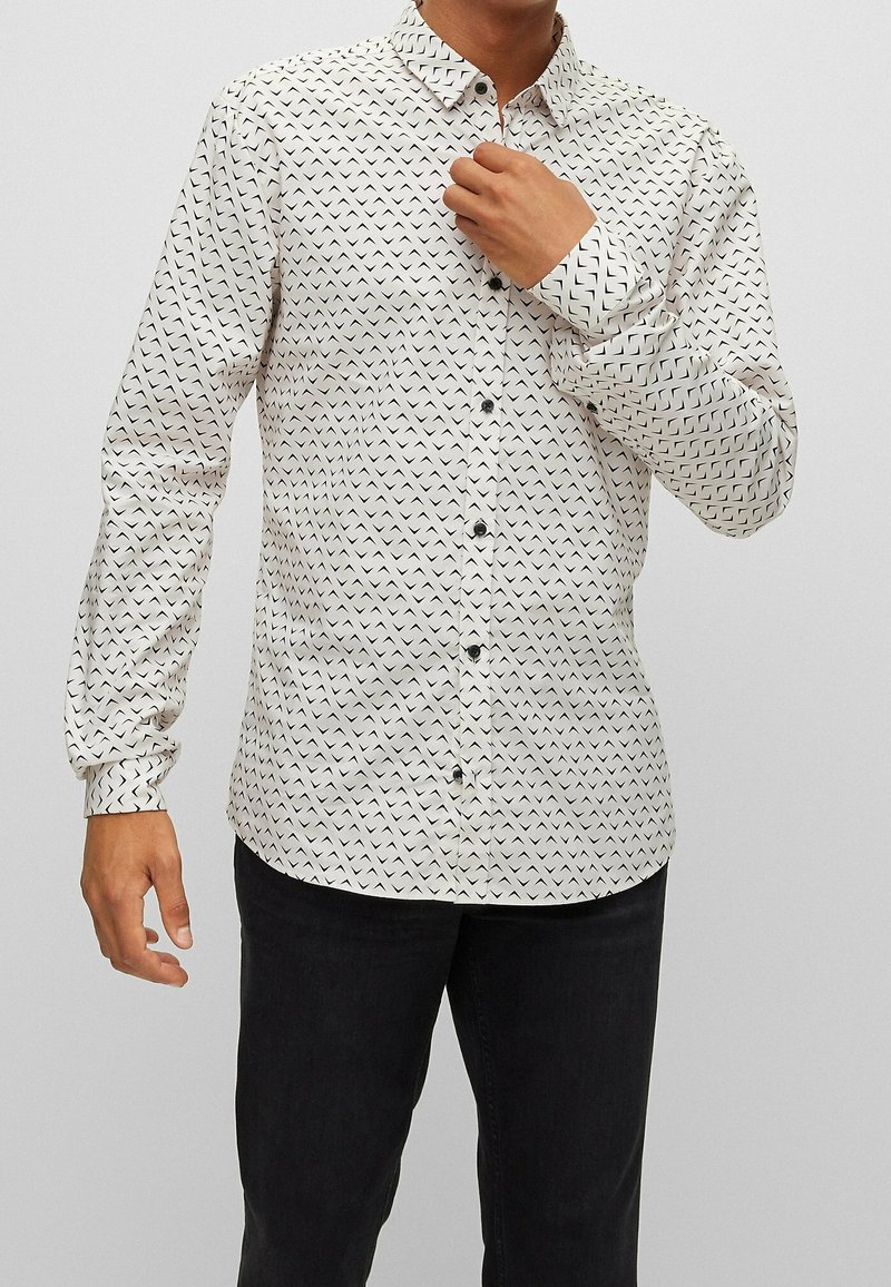 Homme portant une chemise blanche boutonnée avec des motifs géométriques en chevron noirs et un pantalon noir, ajustant le col avec la main droite.