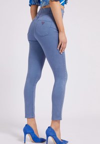 Vrouw in lichtblauwe skinny jeans met hoge taille, een blauw patroon crop top en bijpassende blauwe hoge hakken, staand in profiel.