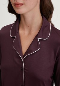 Korte mouwen, donkerpaarse pyjama-top met witte piping langs de kraag en de sluiting, met knoopsluiting en een gladde stoftextuur.