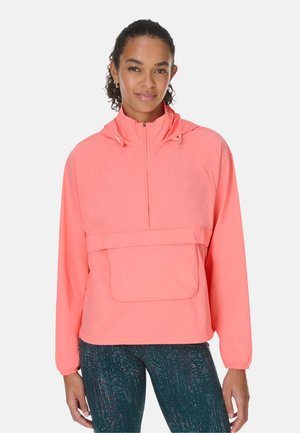 Vrouw in een koraalroze capuchontrui met een voorzak en donkere legging met patroon, staand tegen een effen achtergrond.