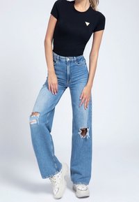 Černé body s krátkým rukávem a trojúhelníkovým logem, doplněné modrými džínami s vysokým pasem, roztrhanými v širokém střihu, a bílými teniskami s potiskem.