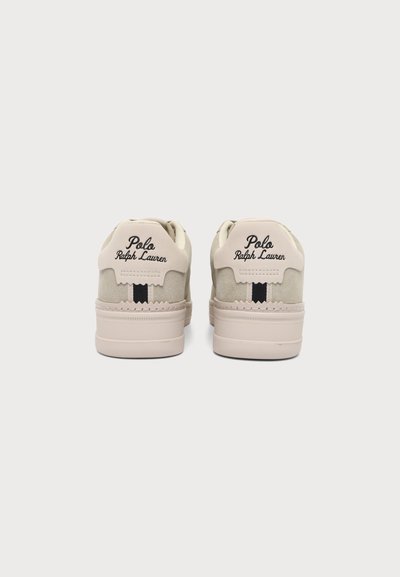 Ζευγάρι μπεζ αθλητικά παπούτσια Polo Ralph Lauren εμφανίζονται από πίσω με μαύρες ριγέ λεπτομέρειες και λογότυπο στο ταμπελάκι της φτέρνας πάνω σε απλό φόντο.