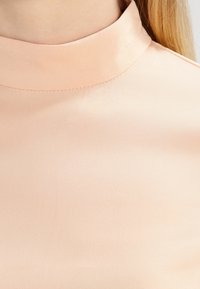 Blusa smanicata in morbida satin color pesca con un collo alto e tondeggiante. Presenta una texture liscia e lucida e un delicato dettaglio di cucitura.