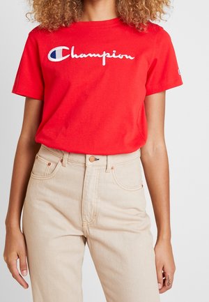 Mujer vistiendo una camiseta roja brillante con la marca Champion, metida dentro de unos vaqueros beige de cintura alta, de pie frente a un fondo liso.