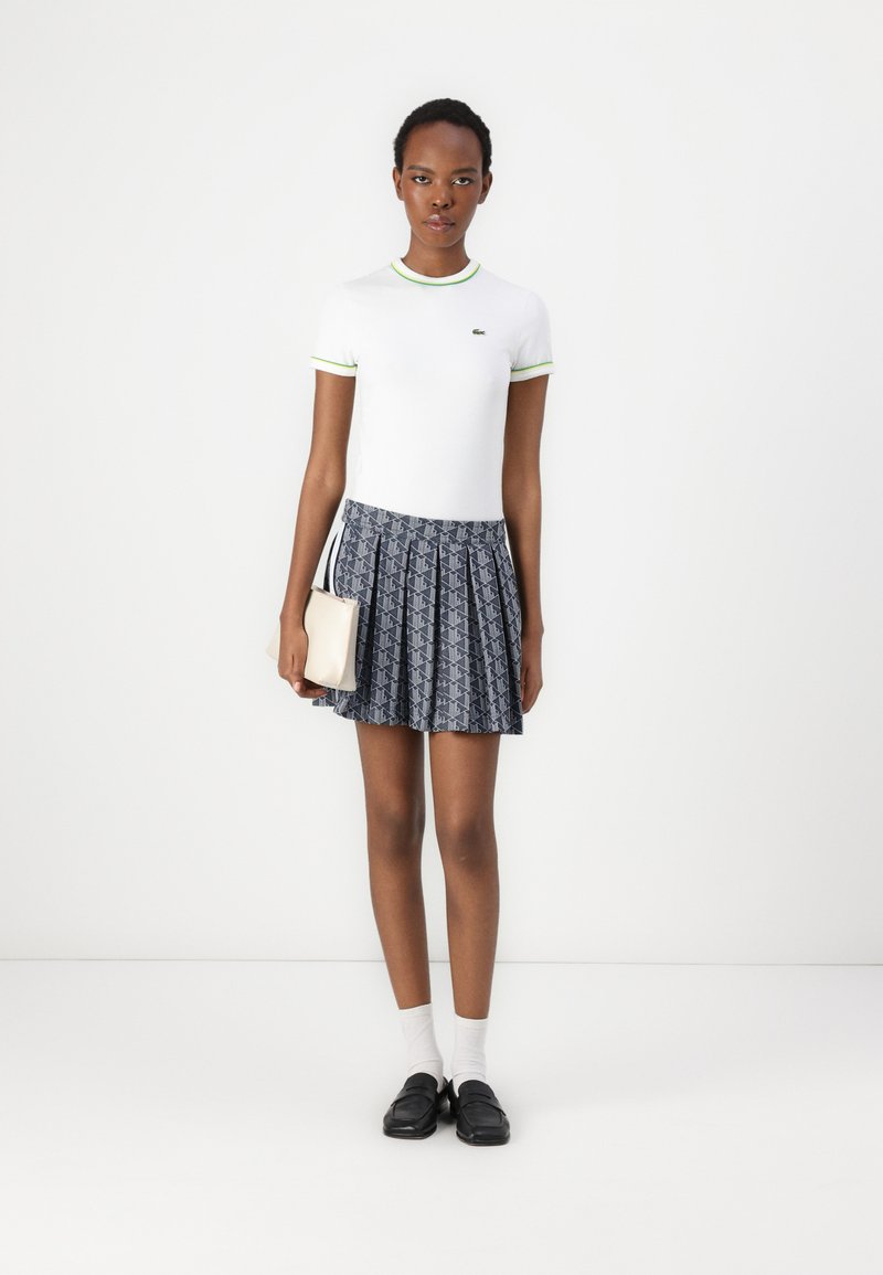 Biała koszula z krótkim rękawem z neonowozielonym wykończeniem, w połączeniu z plisowaną granatową spódnicą z geometrycznym wzorem; trzymając małą beżową torebkę.