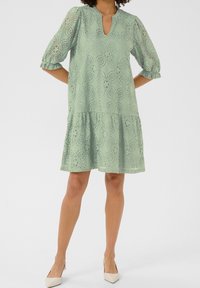 Pitkä hihaton pitsimekko pehmeässä vihreässä sävyssä, jossa on puffihihat ja avonainen pääntie. Kerroksellinen muotoilu kukkakuoseilla ja kevyellä rakenteella.