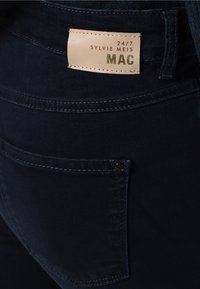 Un bolsillo trasero de unos pantalones vaqueros de mezclilla azul oscuro con una etiqueta de parche de cuero beige que dice "24/7 Sylvie Meis MAC" cosida encima del bolsillo.