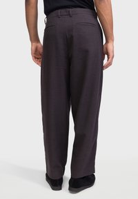 Mann, der dunkelgraue, locker sitzende Hose und schwarze Schuhe trägt, von hinten vor einem schlichten hellen Hintergrund gesehen.