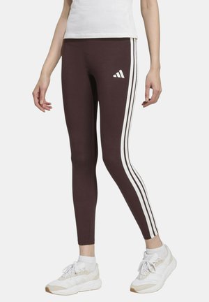Donna che indossa leggings marroni con strisce laterali bianche e logo Adidas, scarpe da ginnastica bianche e top corto bianco, in piedi davanti a uno sfondo bianco.