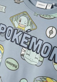 Svetlo modra majica s kapuco z grafiko "POKEMON". Vključuje več ilustracij Pokémonov v različnih oblikah in barvah, z teksturiranim tiskom.