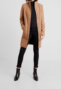 Beige villasekoittunut takki, jossa on vetoketju, korkea kaulus ja sivutaskut. Yhdistetty mustiin farkkuihin ja säärellisiin korkokenkiin.