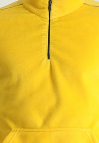 Sudadera de forro polar amarillo con cuello de media cremallera, bolsillo canguro y textura suave. Presenta un detalle de cremallera negra contrastante.