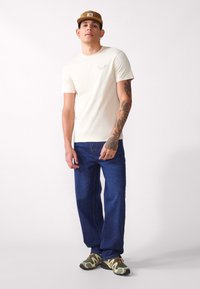 Beige cotton t-shirt with short sleeves and a small graphic on the chest, paired with blue denim jeans and greenish-brown sneakers.