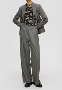 Grau maßgeschneideter Blazer über einem schwarz-weißen gemusterten Top, kombiniert mit hochtaillierten grauen weiten Hosen und schwarzen Slippern.