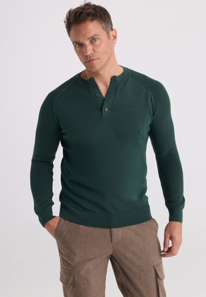 Homme portant un tee-shirt henley à manches longues vert foncé et un pantalon cargo marron, debout avec une main dans la poche sur un fond uni.