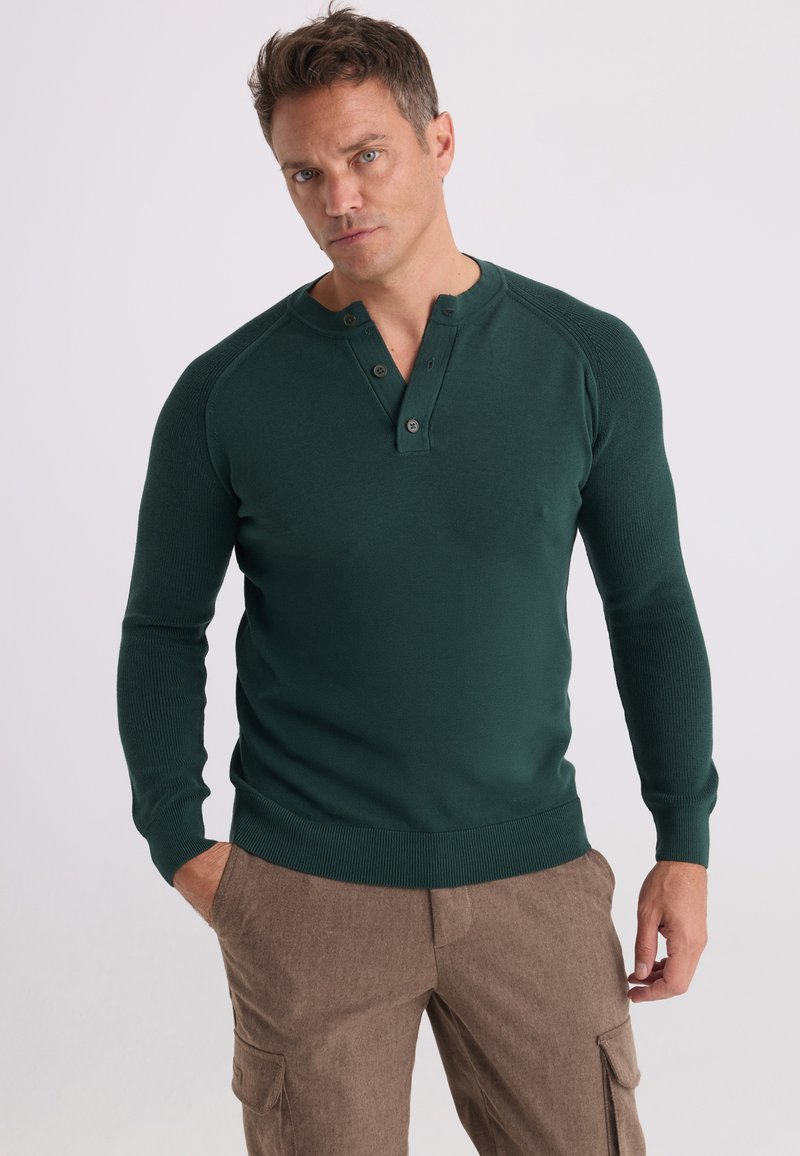 Homme portant un tee-shirt henley à manches longues vert foncé et un pantalon cargo marron, debout avec une main dans la poche sur un fond uni.