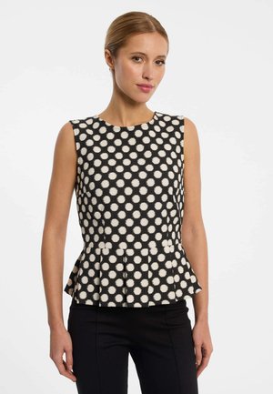 Frau trägt ärmellose schwarze Bluse mit großen weißen Polka-Dots und leichtem Peplum-Saum, kombiniert mit schwarzer Hose, vor weißem Hintergrund.