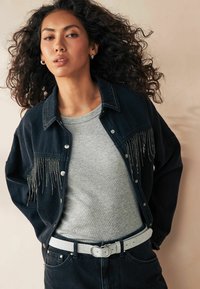 Giubbotto di jeans scuro con dettagli in frange argentate, indossato sopra una t-shirt grigia. Una cintura argentata accentua i jeans scuri, creando un look a strati.