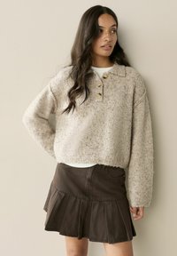 Žena s dlhými tmavými vlasmi, oblečená v béžovom vlnenom svetri s gombíkmi a tmavej plisovanej sukni, stojaca pred jednoduchou svetlou stenou.