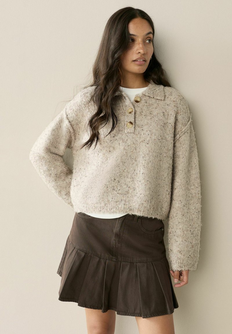 Žena s dlhými tmavými vlasmi, oblečená v béžovom vlnenom svetri s gombíkmi a tmavej plisovanej sukni, stojaca pred jednoduchou svetlou stenou.