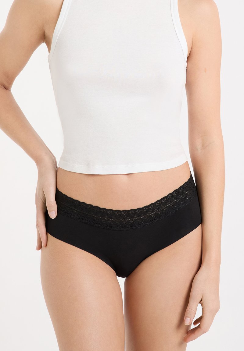 Braguitas negras con una cinturilla de encaje festoneado, que presentan una textura suave y un diseño de cobertura moderada. Camiseta blanca visible por encima.