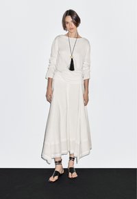 Witte top met lange mouwen gecombineerd met een luchtige witte rok en zwarte sandalen. De rok heeft een gelaagd ontwerp; de top heeft een losse pasvorm en opgerolde mouwen.