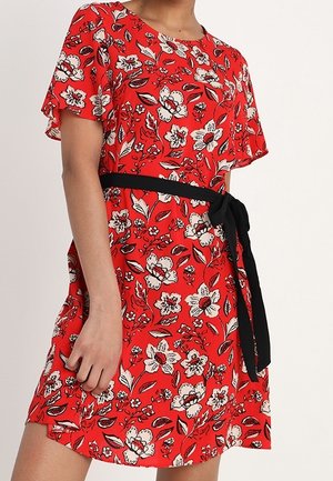 Robe fleurie rouge avec des fleurs blanches et noires, manches courtes et ceinture noire nouée à la taille sur une personne debout.