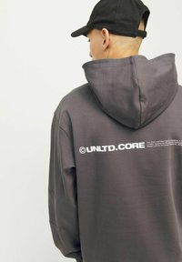 Grijze hoodie met een grote, zichtbare capuchon. Kenmerkt zich door een vet wit logo op de achterkant. Het materiaal lijkt zacht en gestructureerd. Een zwarte pet wordt op het hoofd gedragen.