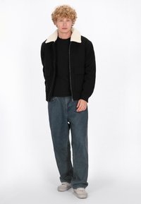 Ung mand med krøllet blond hår står oprejst iført en sort jakke med hvid krave, sort skjorte, løse blå jeans og hvide sneakers.
