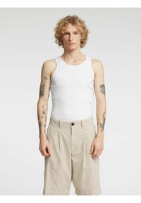 Hvit tanktopp med ribbet tekstur kombinert med beige plisserte shorts med knappelukking. Modellen har krøllete hår og synlige tatoveringer.