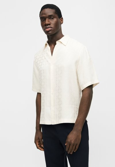 Νέος άνδρας που φοράει κοντομάνικο πουκάμισο σε κρεμ απόχρωση με διακριτικό μοτίβο, συνδυασμένο με σκούρο παντελόνι, στέκεται μπροστά σε απλό φόντο.