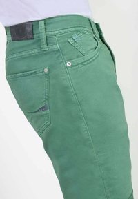 Groene spijkerbroek heeft een fitted ontwerp, rechte pijpen en een kenmerkende achterzak met een klein label. De stof heeft een gladde textuur.