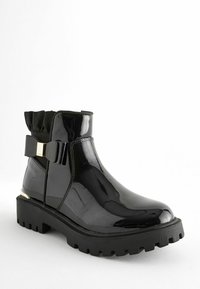 Botas de tobillo de charol negro con una suela gruesa y texturizada, parte superior de textil elástico y un lazo decorativo con un detalle dorado en el lateral.